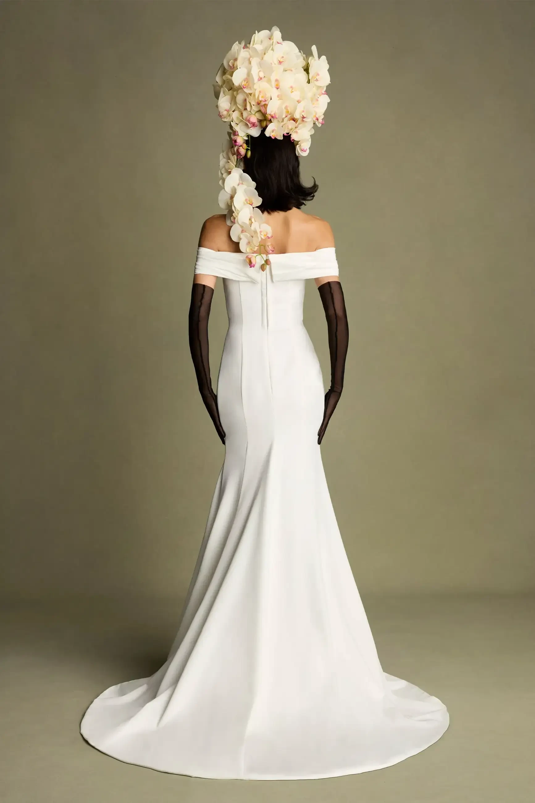 A woman in a white off-the-shoulder gown with a long train, viewed from behind, wearing black sheer gloves and a large floral headdress.