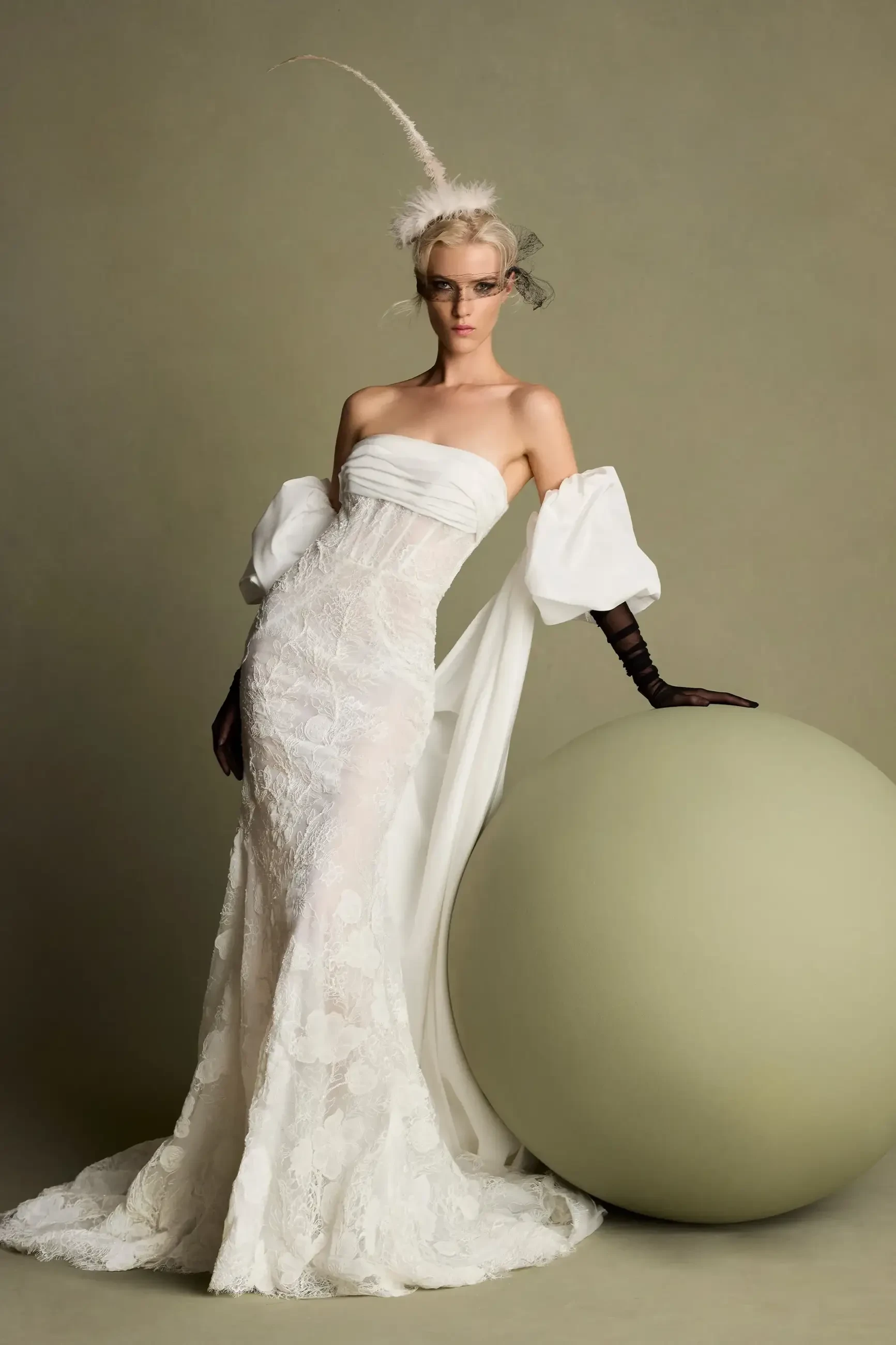 A model wearing an elegant white gown with off-the-shoulder sleeves, standing beside a large, round, pale green object. The gown features intricate detailing and the model has styled hair with a feather accessory.