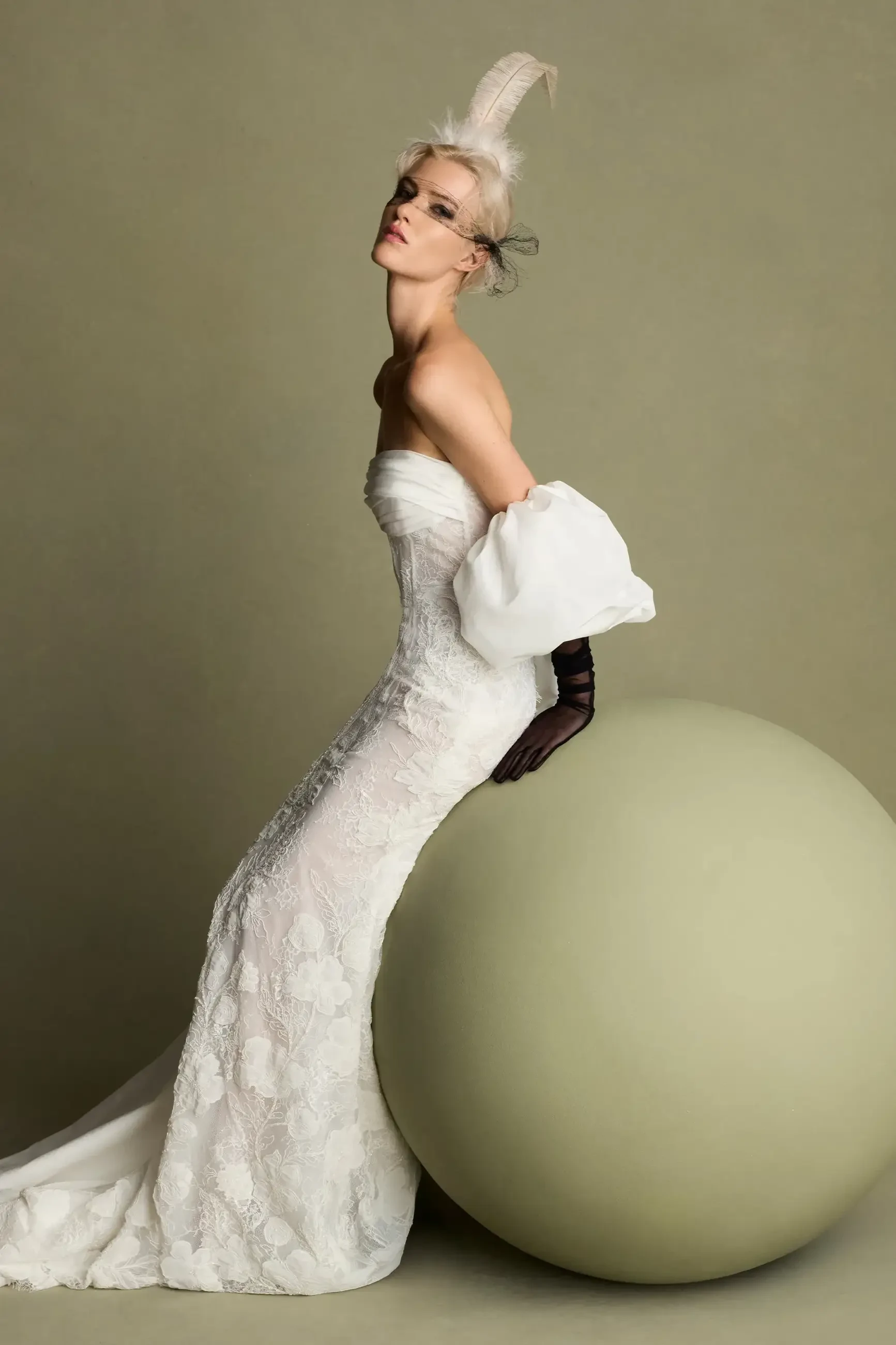A model in a white strapless gown poses elegantly next to a large, smooth, beige sphere, with dramatic makeup and a unique hairstyle.