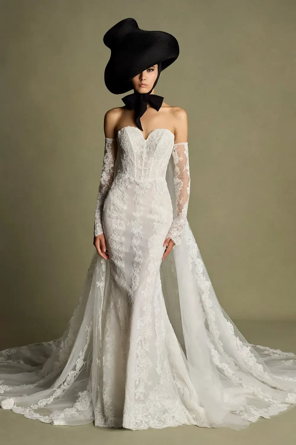 A model wearing an elegant white lace wedding gown with long sleeves and a dramatic train, complemented by a large black floppy hat and a black ribbon.