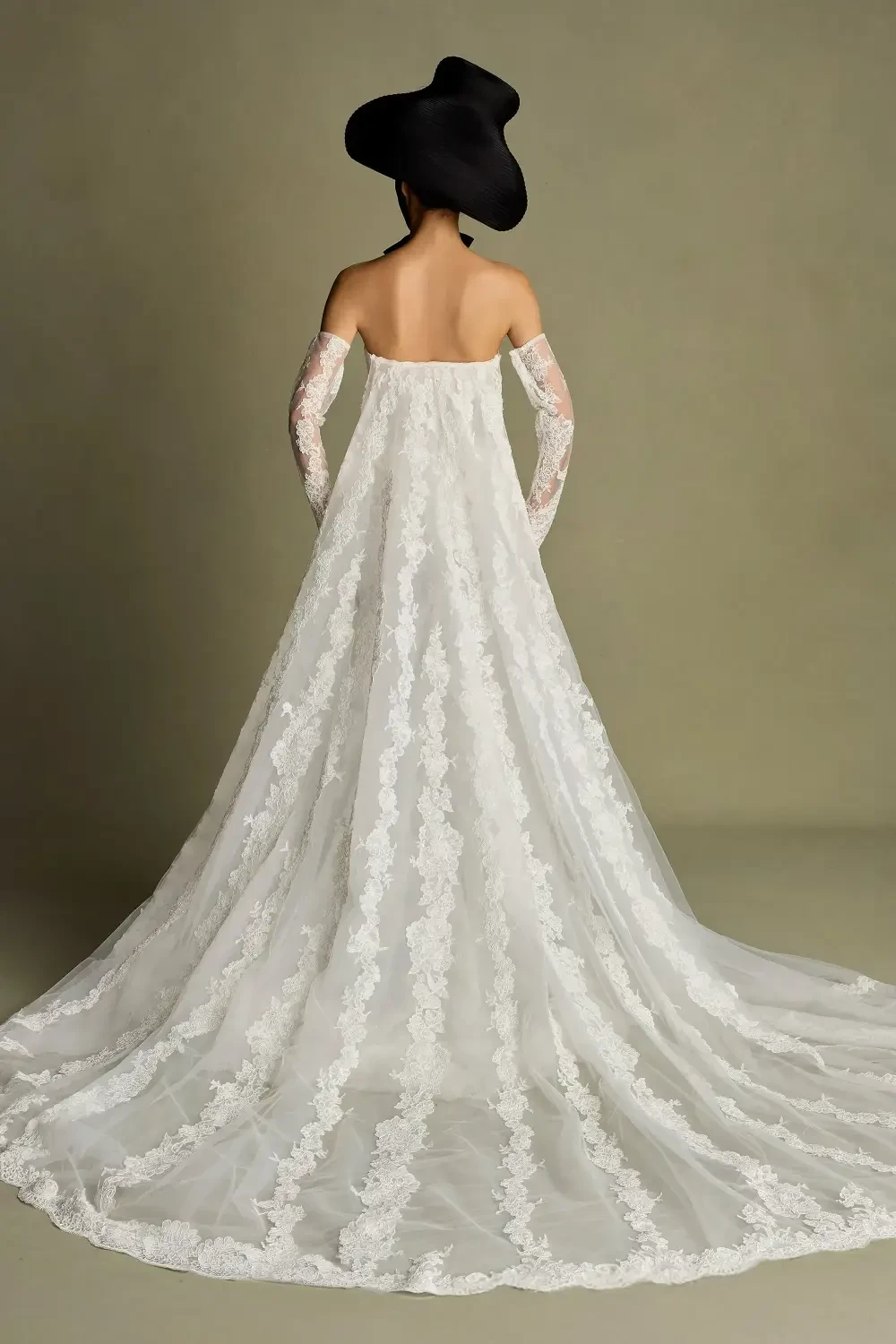A woman in a strapless white gown with a long train and lace detailing, wearing gloves and a large black hat, viewed from behind.