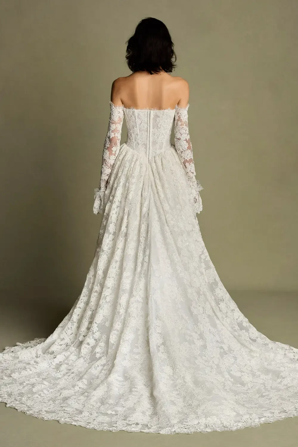 A woman in a white lace wedding dress with long, off-the-shoulder sleeves, viewed from the back. The dress features intricate lace detailing and a flowing train.