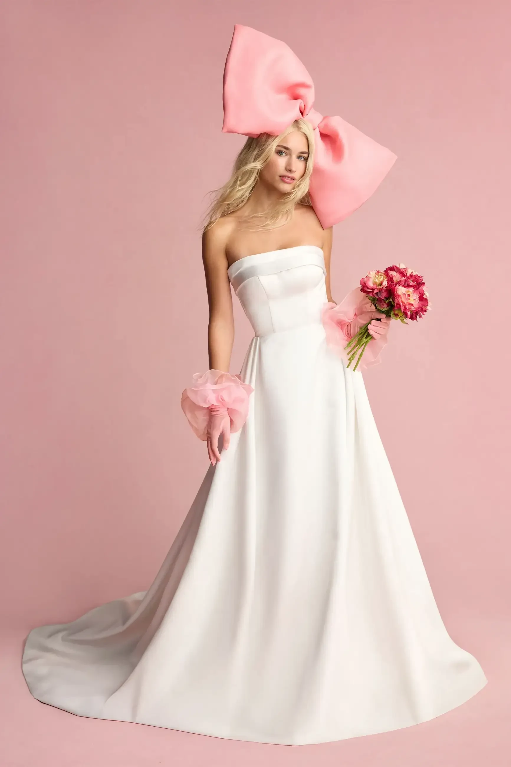 A model wearing a strapless white gown with a large pink bow on her head, holding pink flowers and wearing pink accessories.