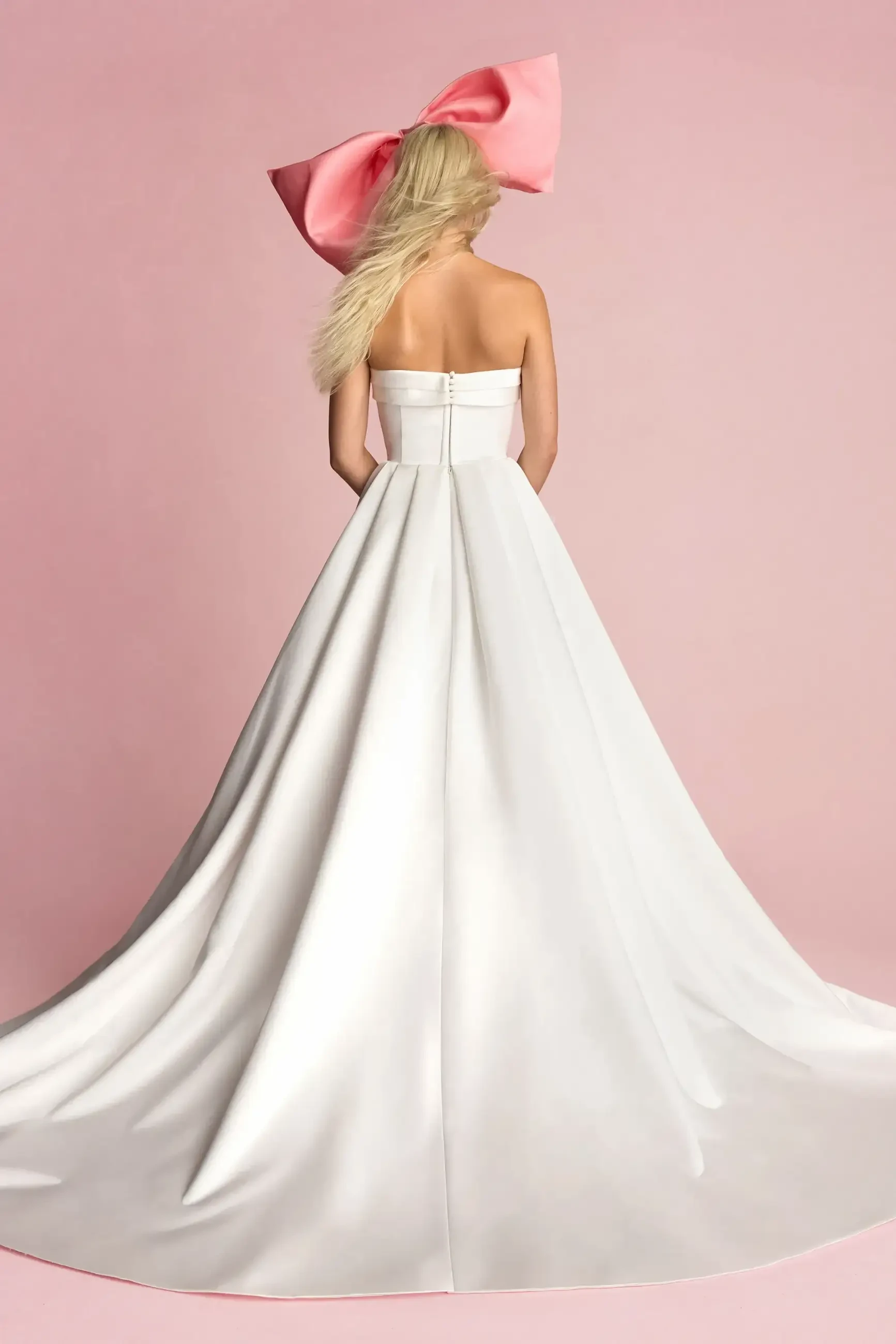 A model is seen from the back wearing a strapless white gown with a long train, and a large pink bow in her hair against a pink background.