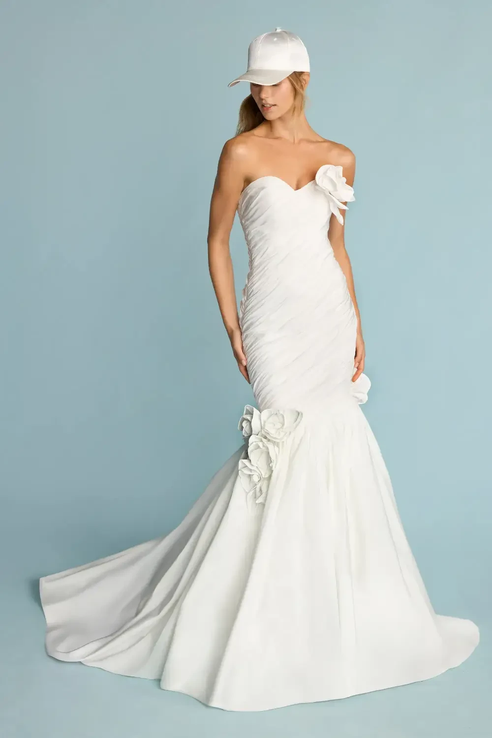 A model wearing a strapless white gown with a mermaid silhouette, featuring floral embellishments, paired with a white baseball cap against a light blue background.