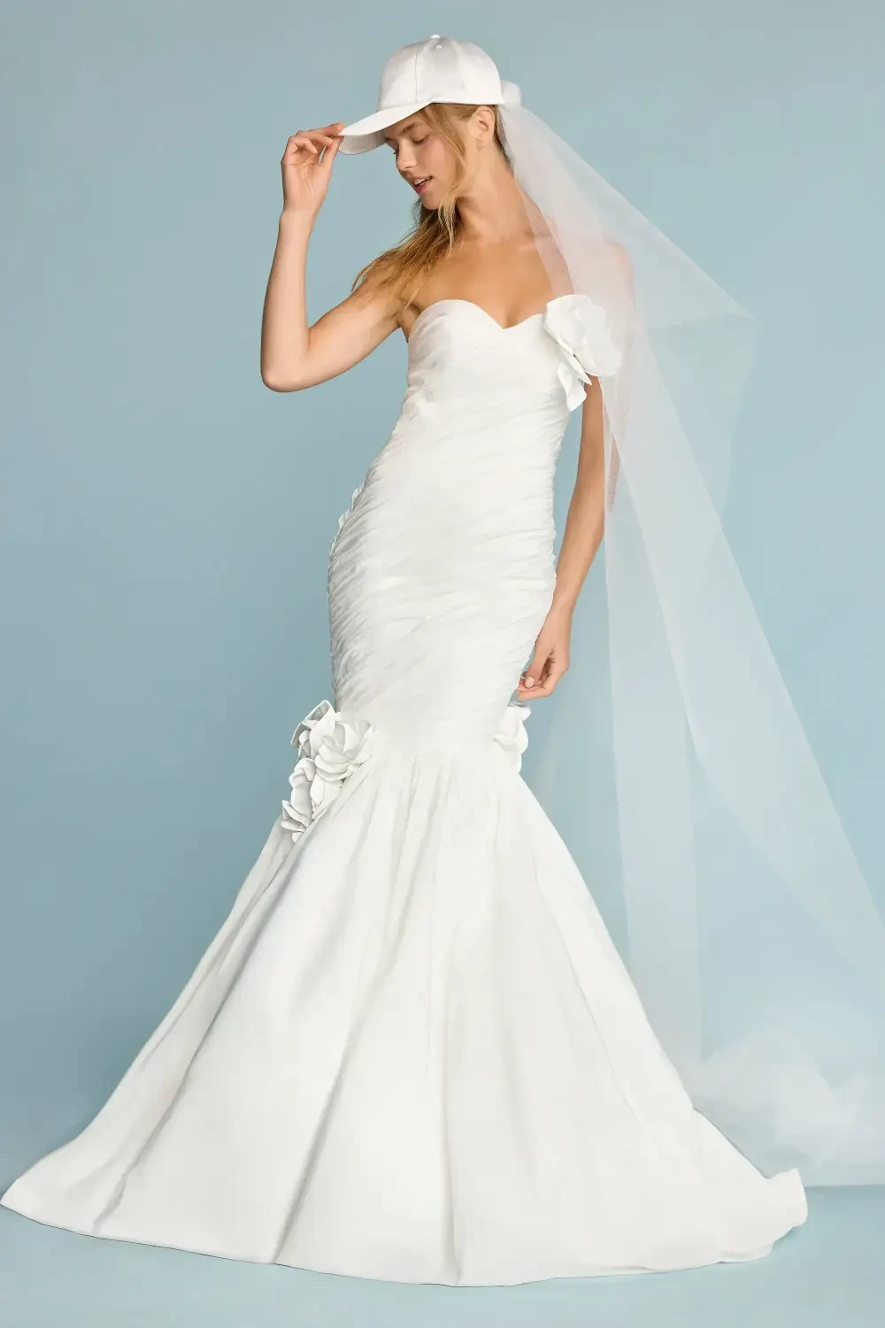 A model wearing a strapless white gown with a mermaid silhouette, featuring floral embellishments, paired with a white baseball cap against a light blue background.