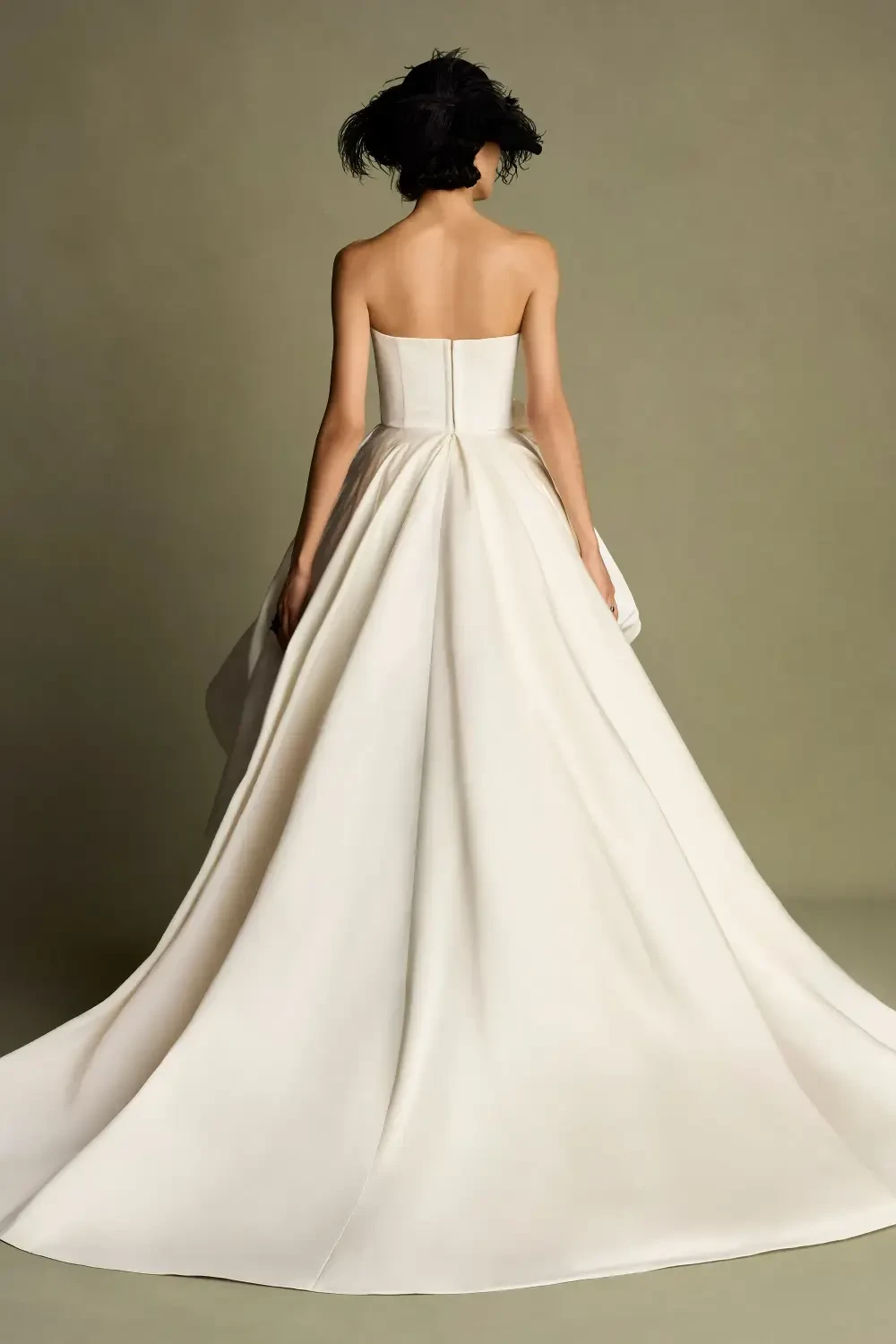 A woman in a strapless white gown with a full train, standing with her back to the camera against a neutral background.