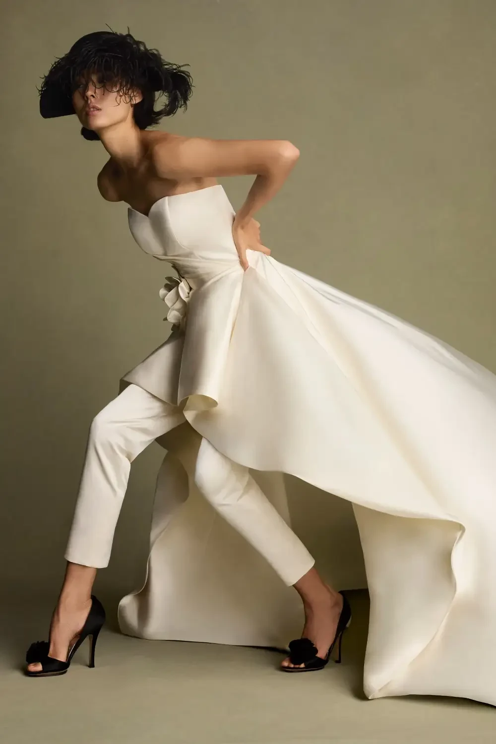 A model wearing a strapless, flowing white gown over fitted pants, striking a dynamic pose against a neutral background.