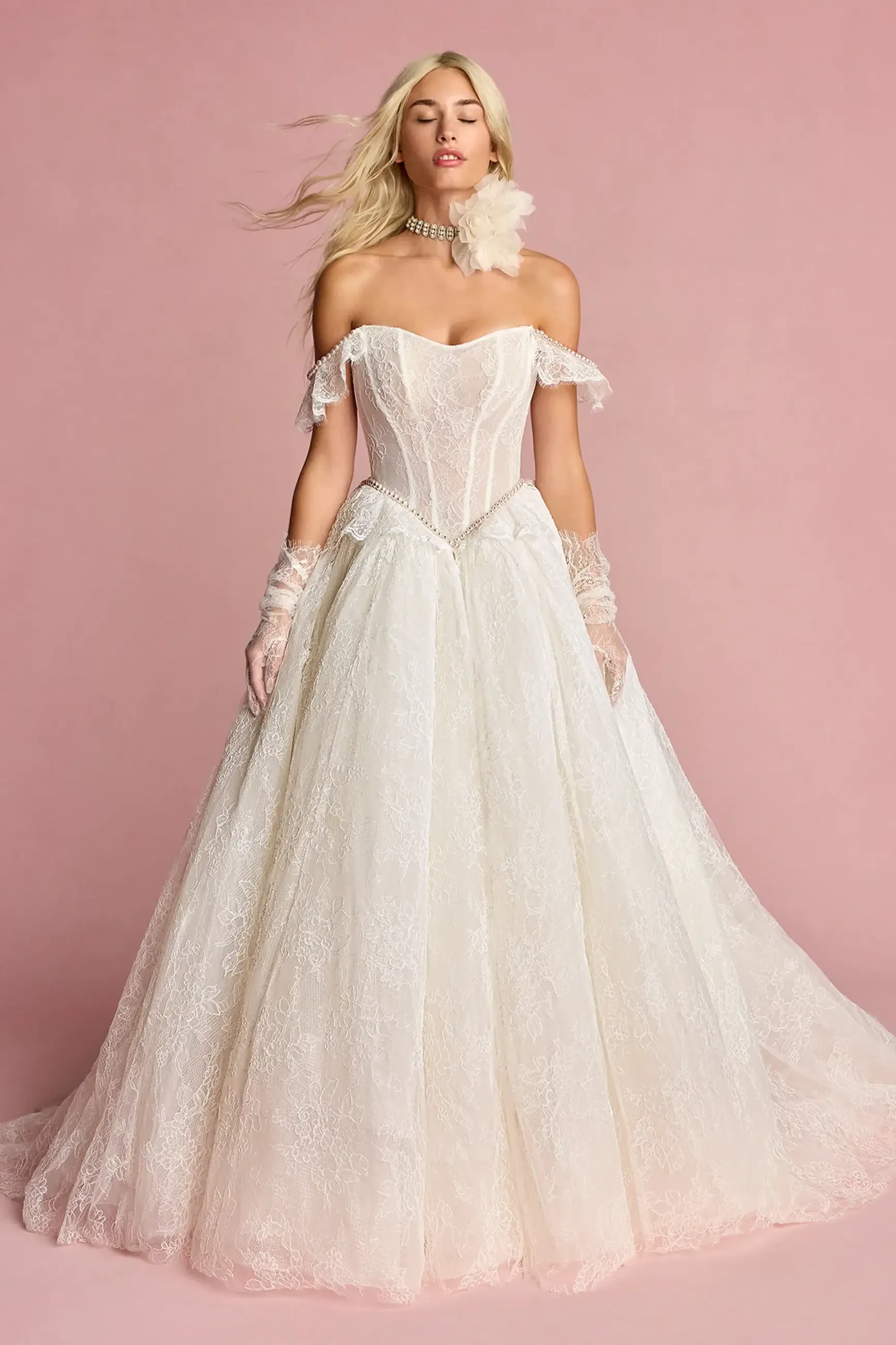 A woman modeling a vintage-inspired off-the-shoulder wedding gown with a full skirt and intricate details, standing against a pink background.