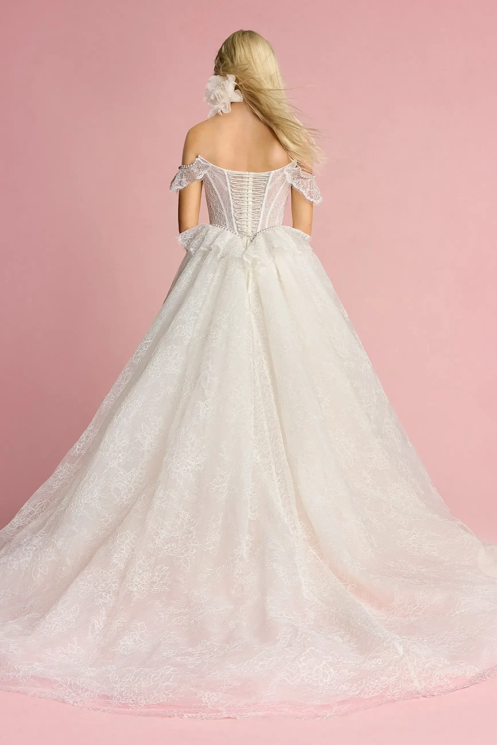 A woman wearing a white wedding gown with an off-the-shoulder design, standing with her back to the camera against a pink background.
